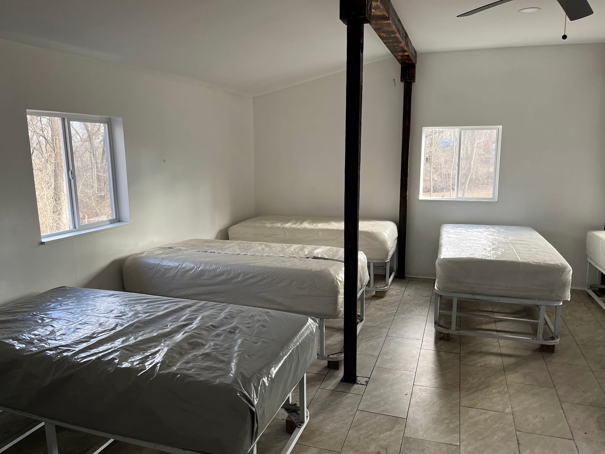 A dormitory-style room with four mattresses on white metal bed frames, still in plastic wrap. Each wall has a small window in it. A dark structural beam runs floor to ceiling in the center of the room.